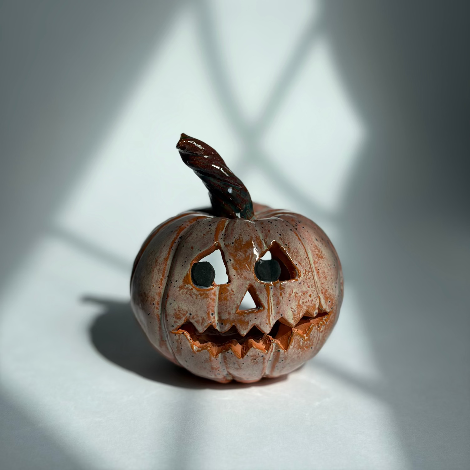 A small carved jack-o'-lantern pumpkin with a dark stem, photographed on a white surface with dramatic window-pane shadows cast across the scene.