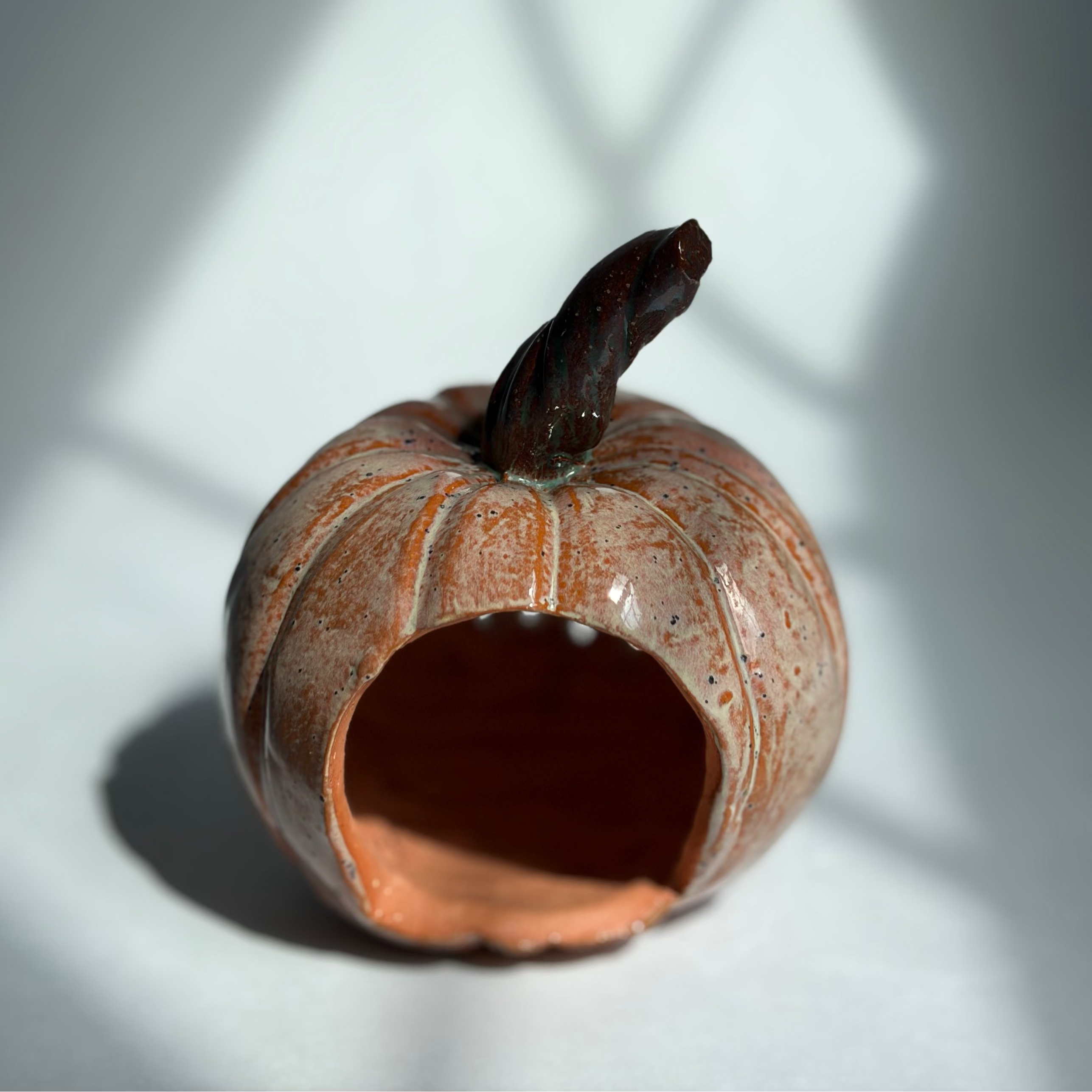 A small decorative ceramic pumpkin-shaped object with a broken open bottom and a dark stem, photographed on a white surface with strong window-shadow lighting.