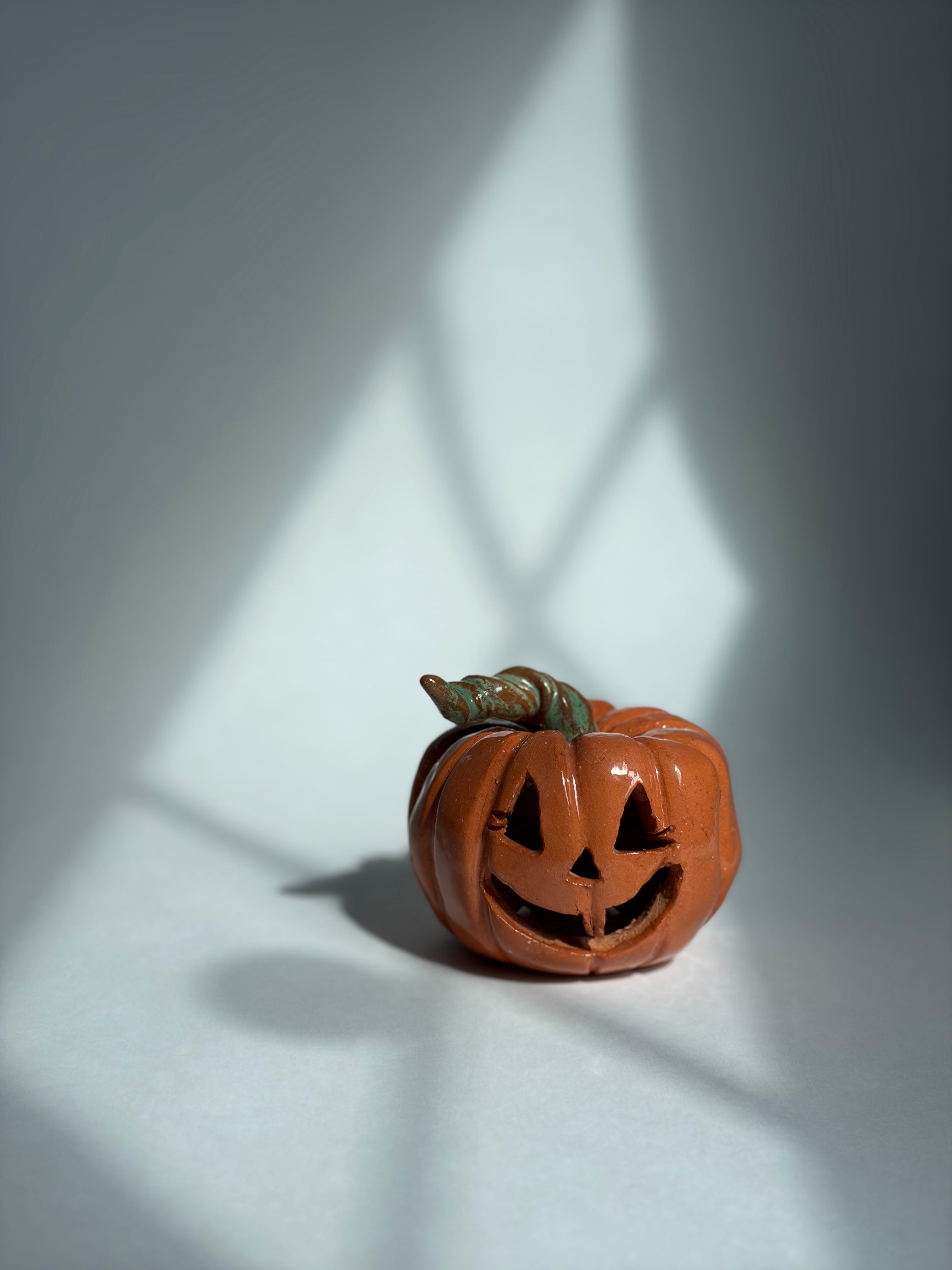 A small carved orange jack-o'-lantern figurine with a green stem, photographed on a white surface with dramatic window-box shadows cast across the scene.