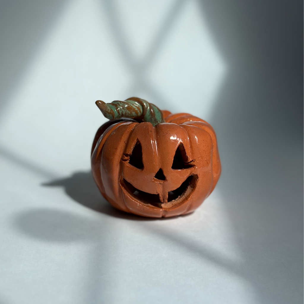 A small carved orange jack-o'-lantern pumpkin figurine with a green stem, photographed on a white surface with dramatic window-light shadows cast across the scene.