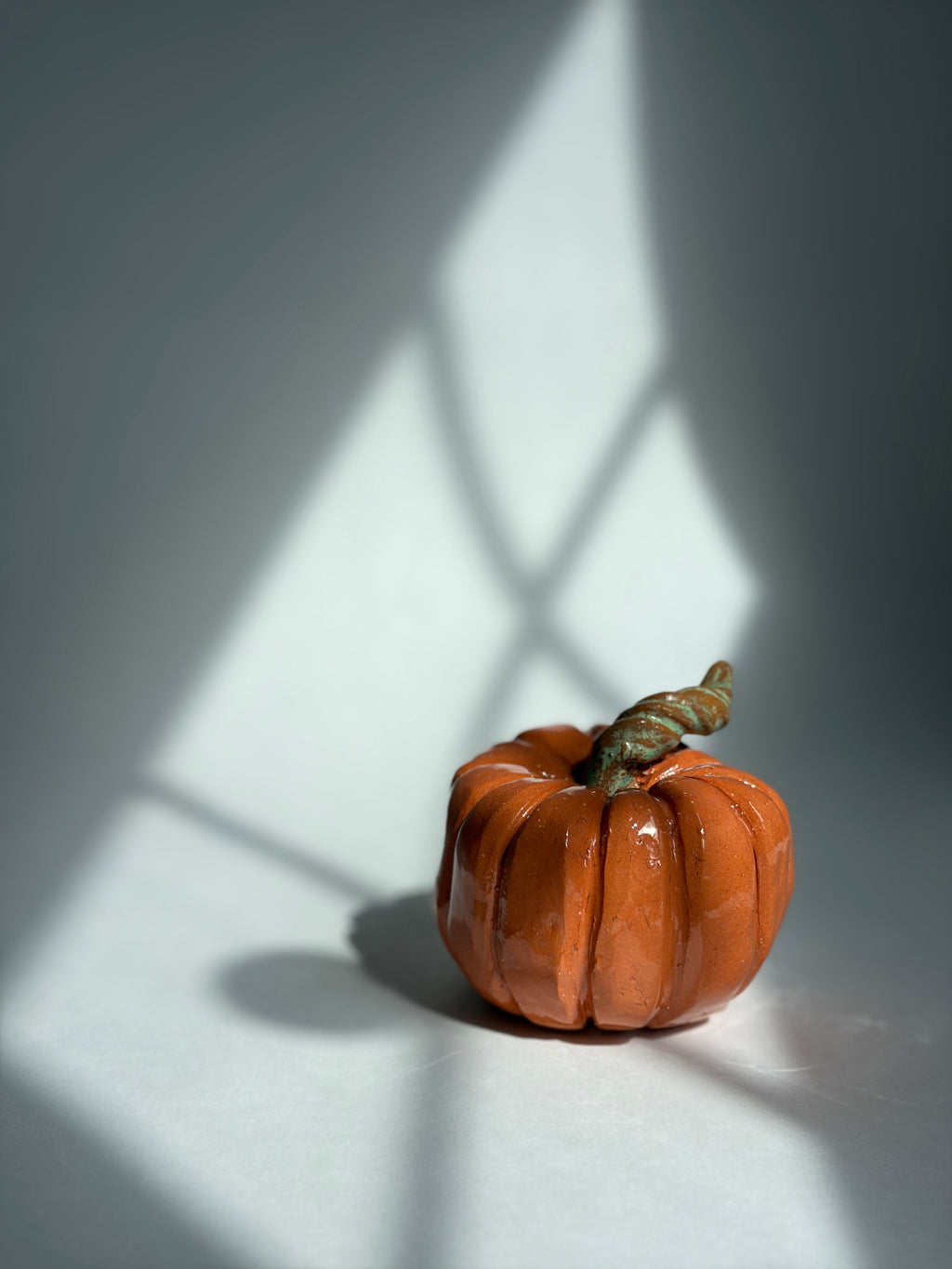 A small orange pumpkin figurine with a textured surface and a brown stem, placed on a white surface with dramatic window-pane shadows cast across it.