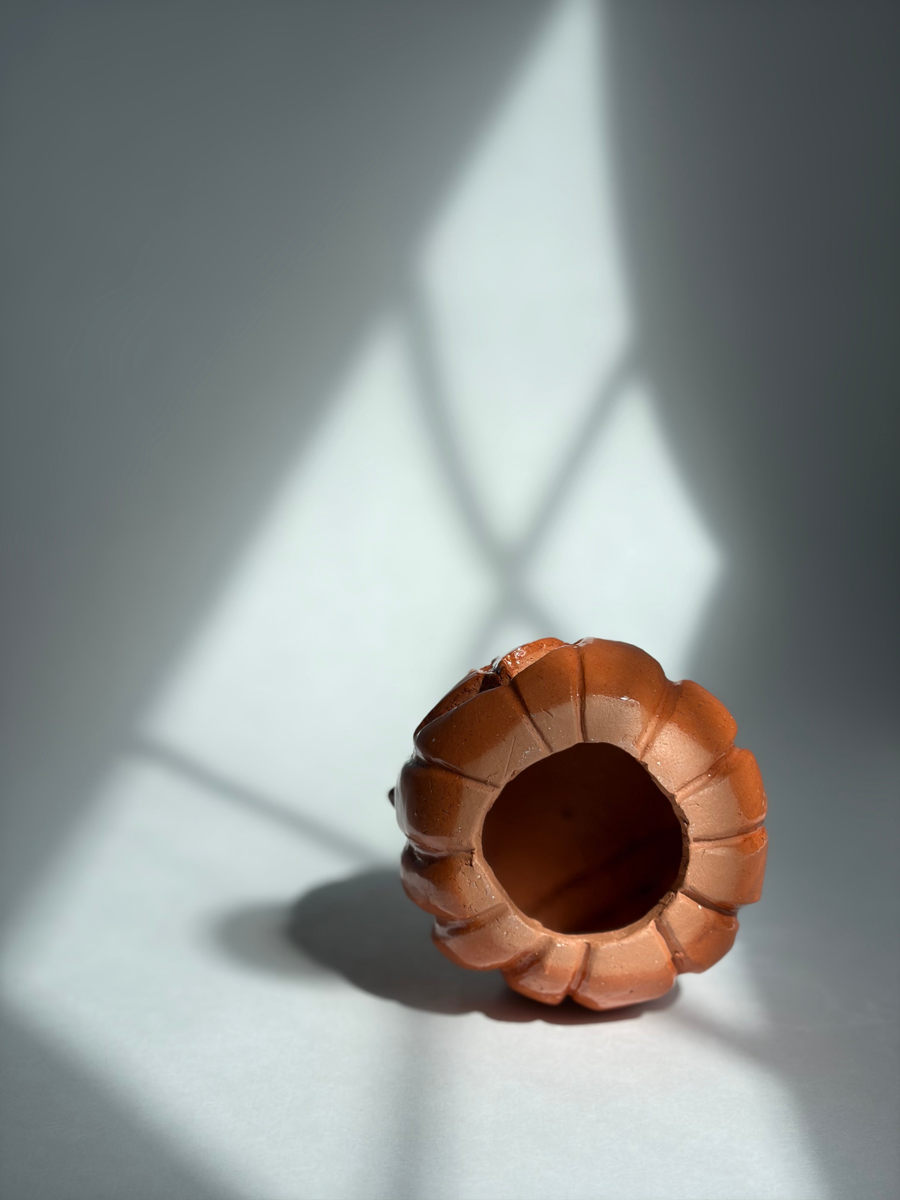 A terracotta-colored ceramic cup with a textured, ribbed exterior and a smooth, open center, photographed on a light gray surface with dramatic diagonal window-light shadows.