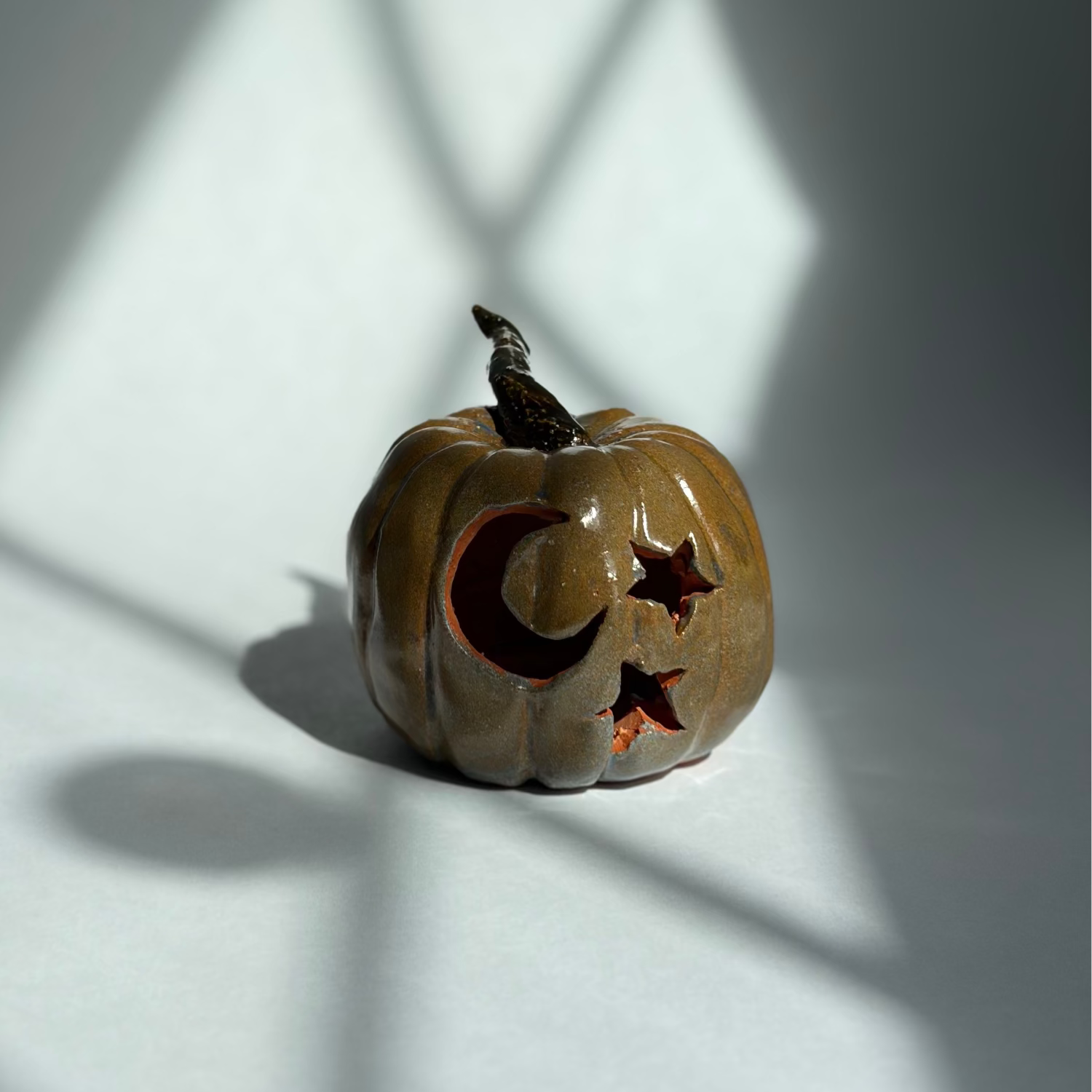 A small carved jack-o'-lantern pumpkin figurine with a glossy brown finish, featuring a crescent moon and two stars, placed on a white surface with dramatic window-light shadows.