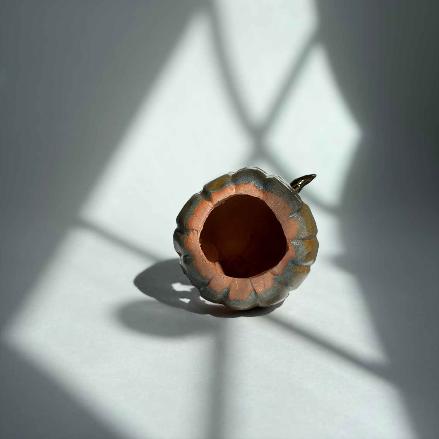 A small decorative ceramic vase shaped like a pumpkin, featuring a warm terracotta orange interior and a gray-blue glazed exterior, set against a white surface with dramatic window shadow patterns.