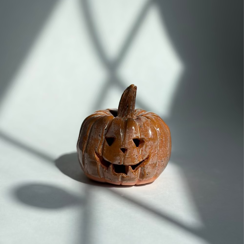 A small carved pumpkin figurine with a carved jack-o'-lantern face sits on a white surface under dramatic window shadow patterns.