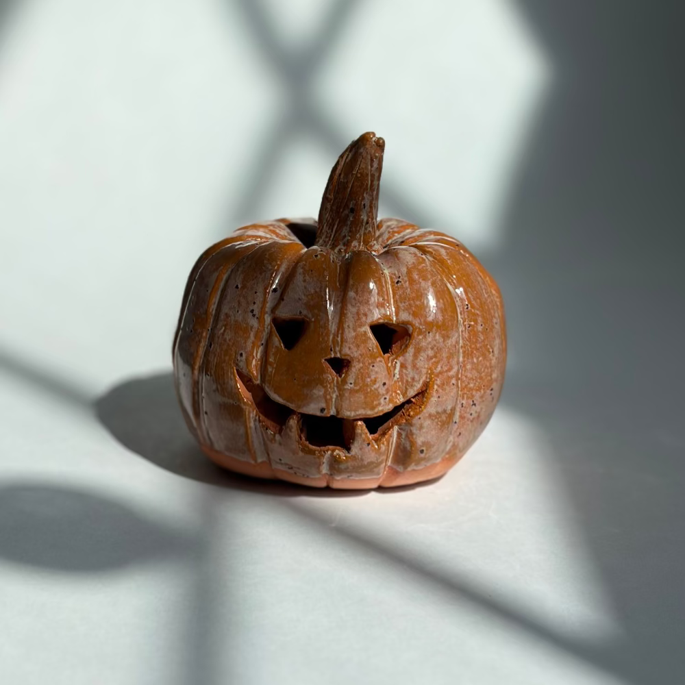 A small carved pumpkin figurine with a carved jack-o'-lantern face sits on a white surface in sharp sunlight, casting a shadow on a gray wall with windowpane shadows.