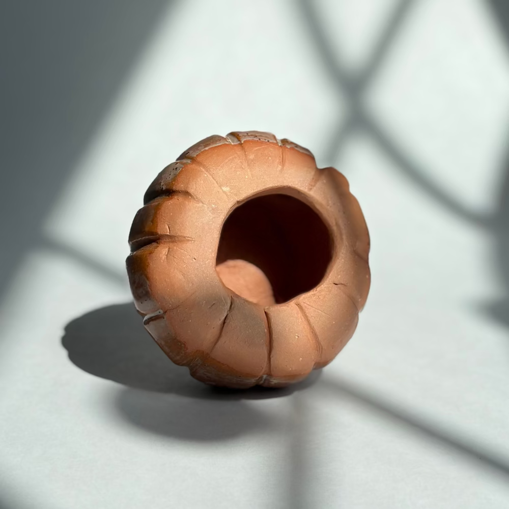 A small sculptural ceramic vase with a textured, ribbed exterior and a hollow center, photographed on a gray surface with dramatic window-light shadows.