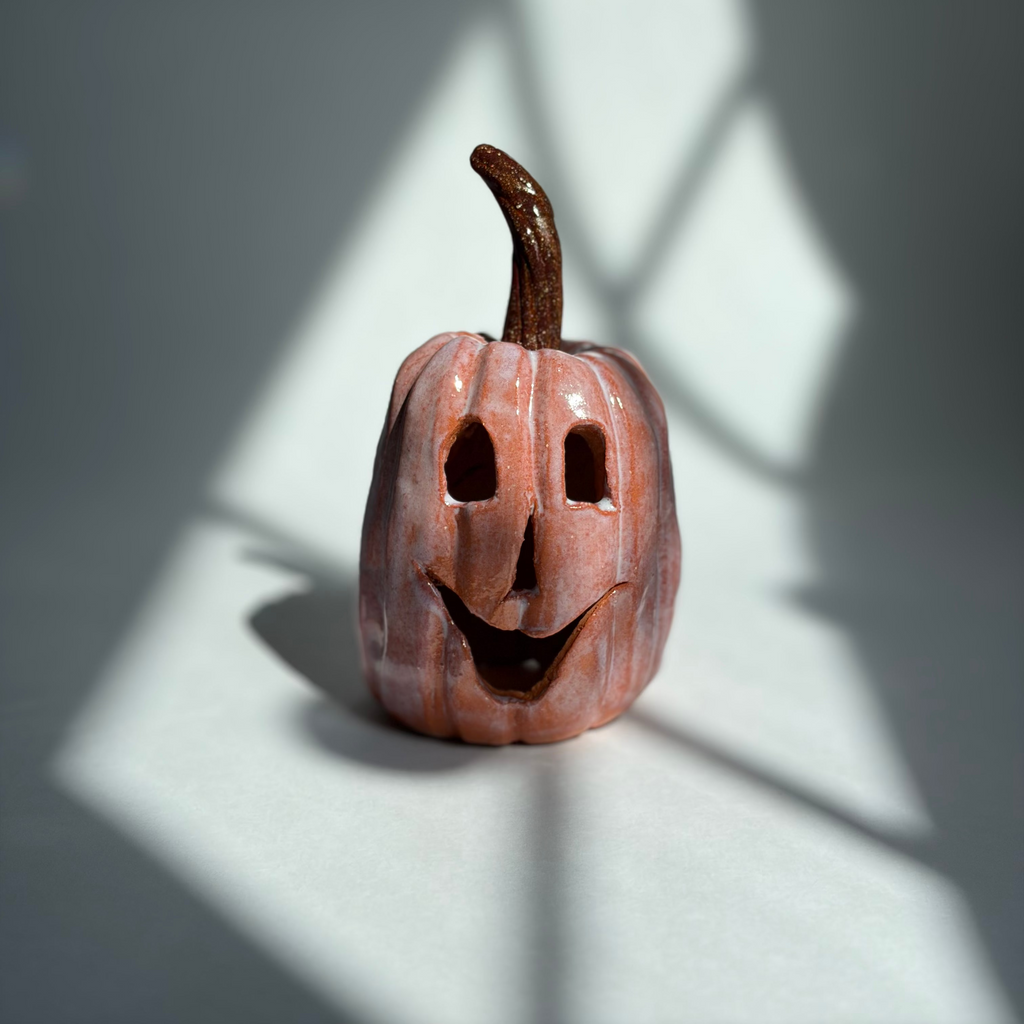 A small carved jack-o'-lantern pumpkin figurine with a dark brown stem, standing on a white surface under dramatic diagonal window shadows.