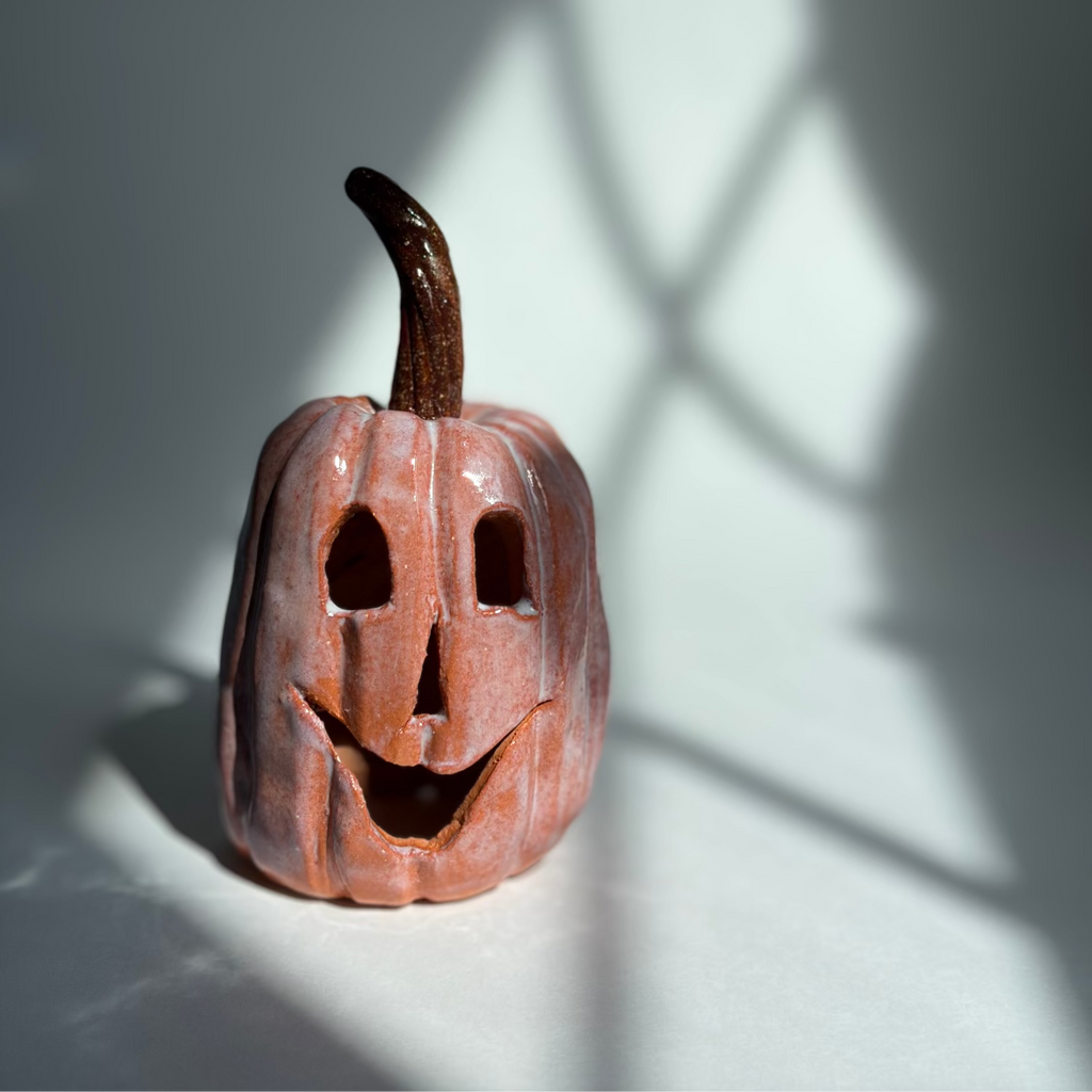 A small carved jack-o'-lantern pumpkin figurine with a glossy terracotta finish and a dark brown stem, photographed on a white surface with dramatic window-shadow lighting.