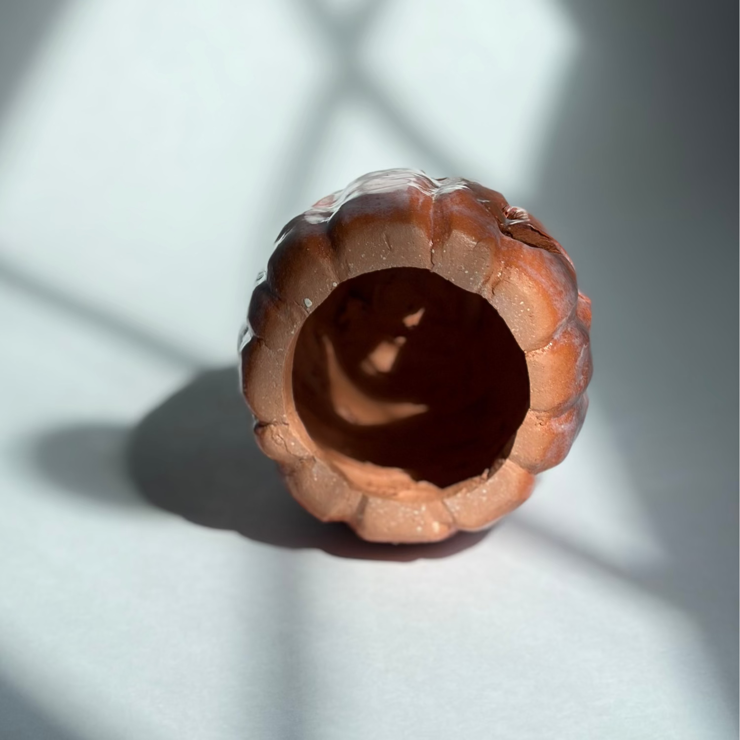 A small carved wooden pumpkin or jack-o'-lantern with a hollow center, photographed on a white surface with dramatic window-shadow lighting.