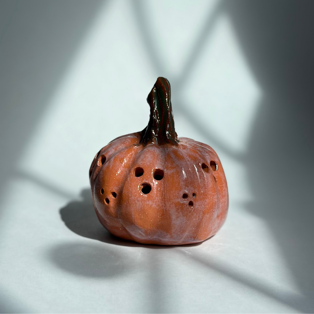 A small decorative orange pumpkin sculpture with a dark stem and several small dark holes on its surface, photographed on a white surface with dramatic window-light shadows cast across the scene.