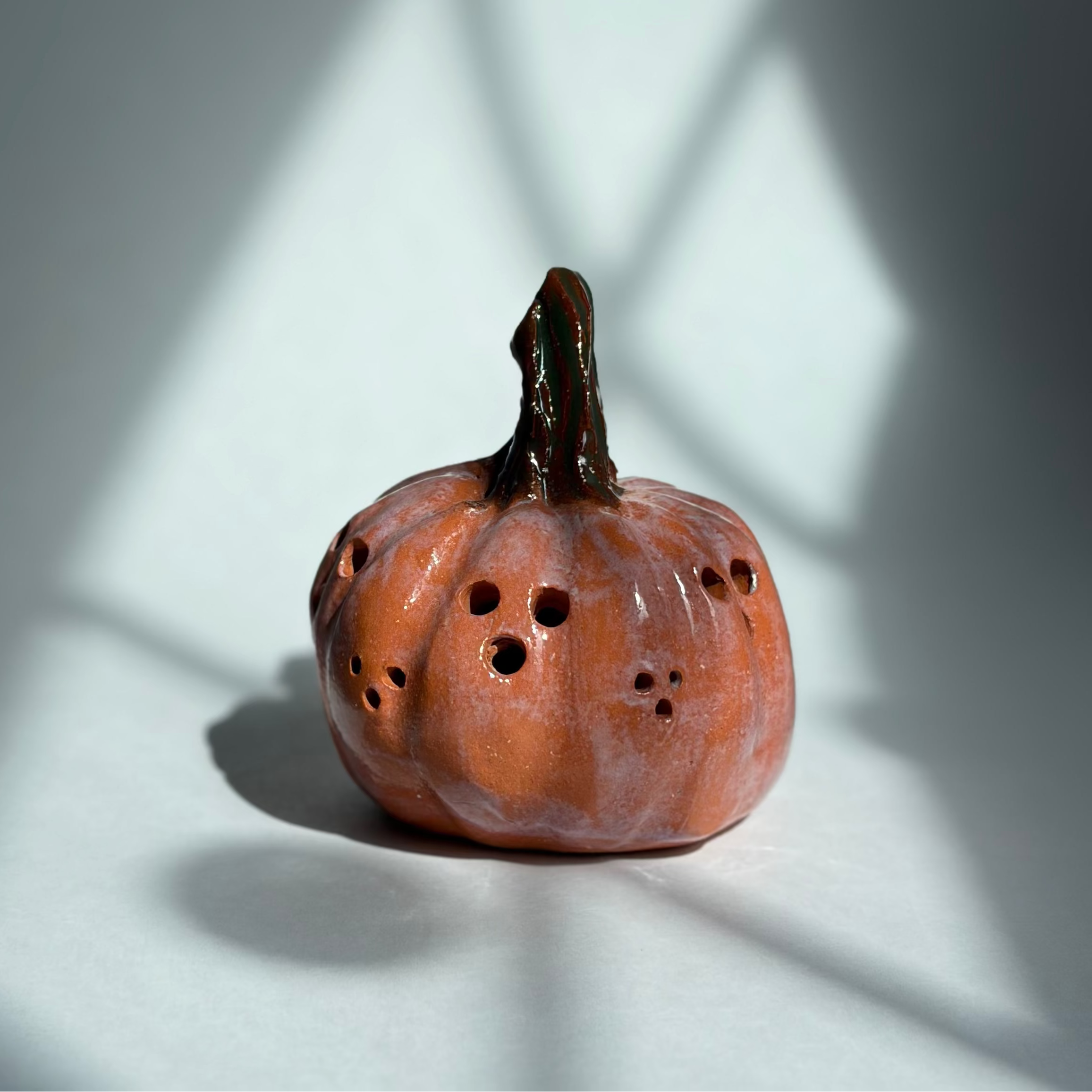 A small decorative orange pumpkin sculpture with a dark stem and several small dark holes on its surface, photographed on a white surface with dramatic window-light shadows cast across the scene.