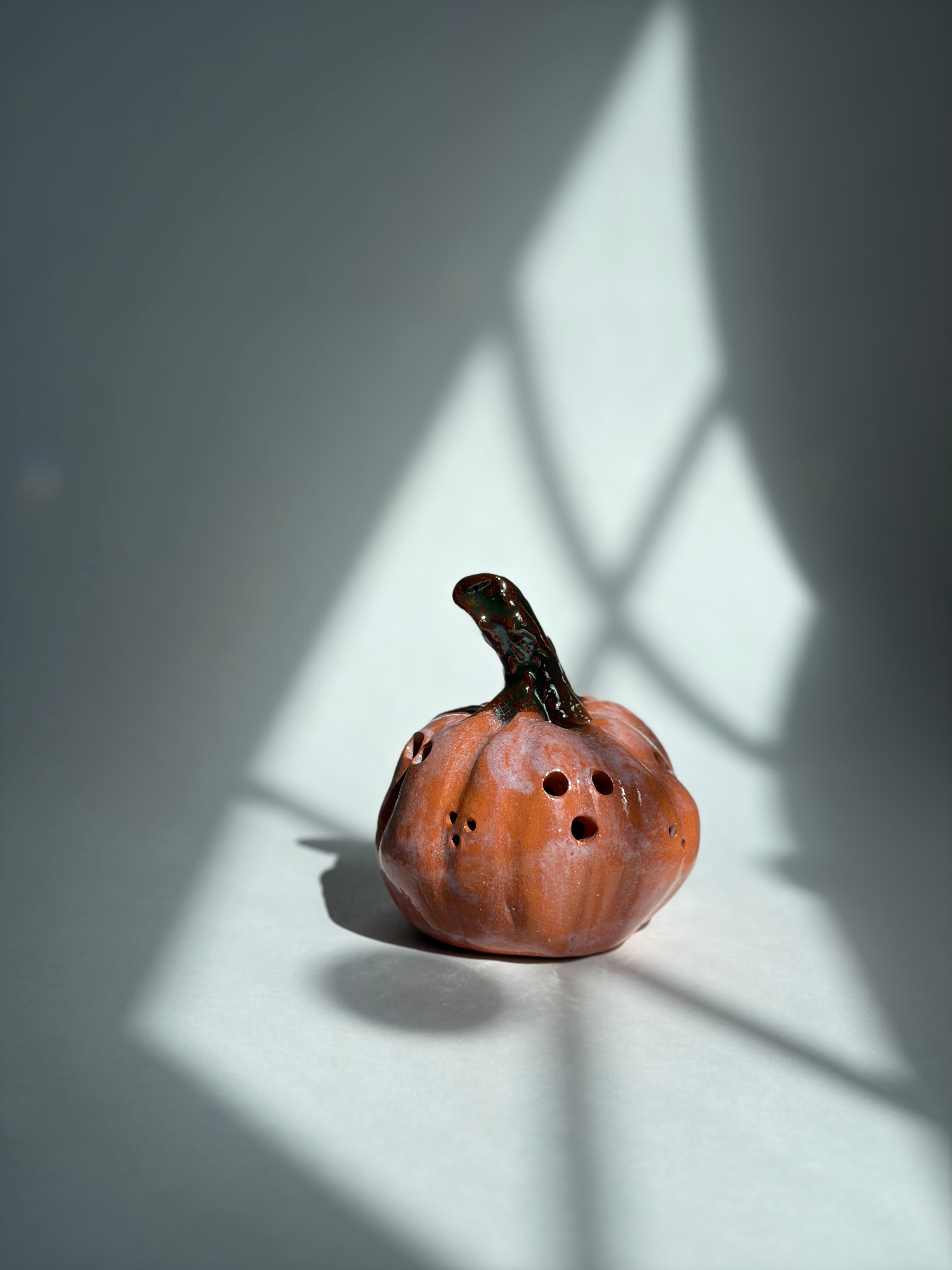 A small carved orange pumpkin figurine with a dark stem and several circular cutouts, photographed on a white surface with dramatic window-shadow lighting casting geometric patterns across the scene.