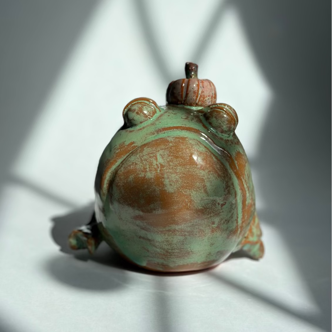 A small ceramic frog figurine with a glossy green and terracotta glaze, shown from the back on a white surface with dramatic diagonal window light shadows.