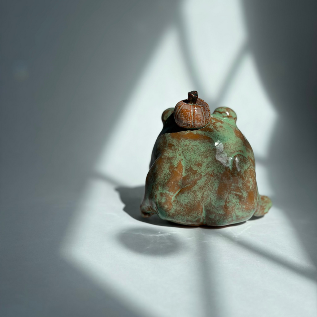 A small ceramic frog figurine with a green and brown patina finish, sitting on a white surface under dramatic window shadow light.