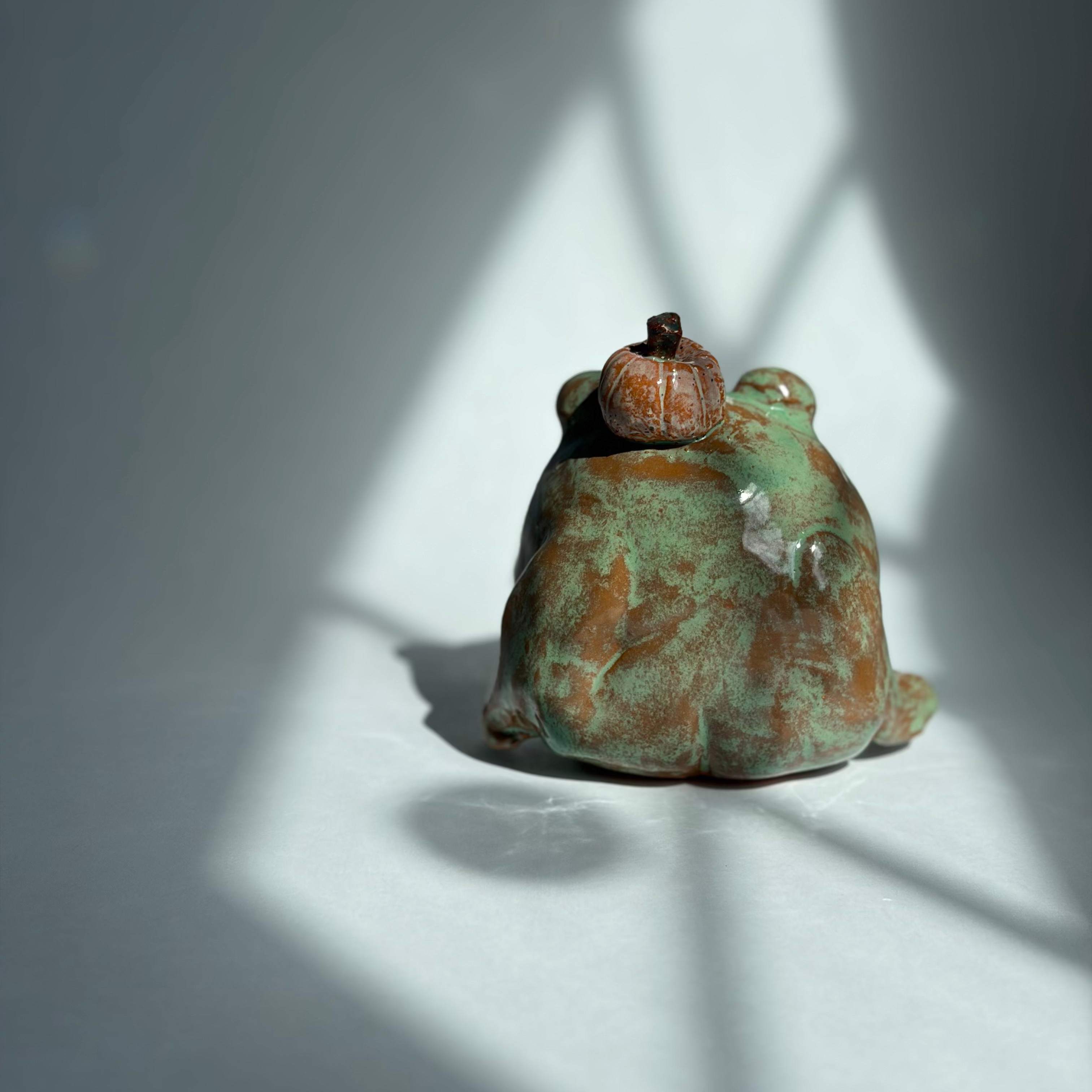 A small ceramic frog figurine with a green and brown patina finish, sitting on a white surface under dramatic window shadow light.