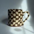 A ceramic mug with a brown and cream checkerboard 3D cube pattern, photographed in dramatic side lighting with strong shadows on a gray background.