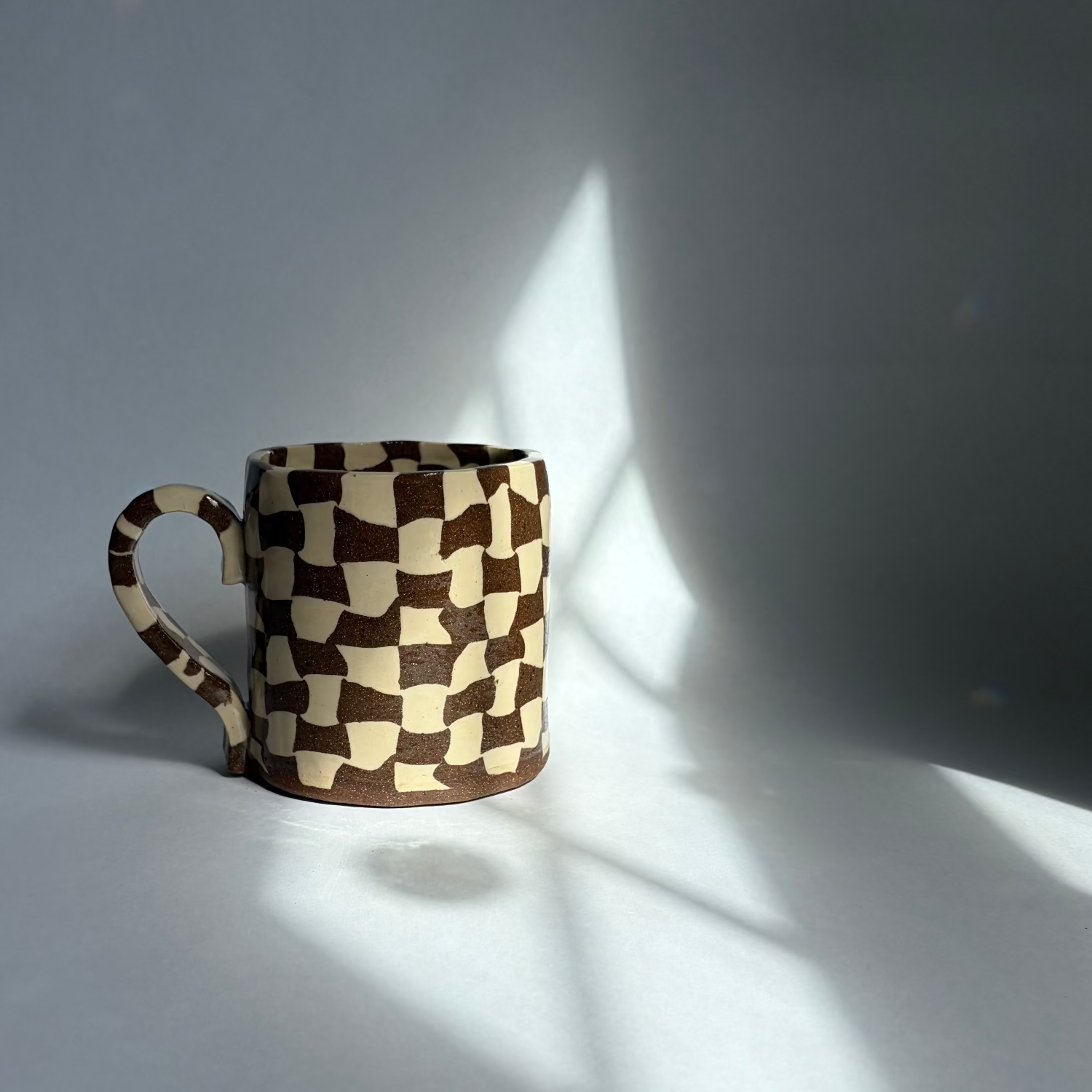 A ceramic mug with a brown and cream checkerboard 3D cube pattern, photographed in dramatic side lighting with strong shadows on a gray background.