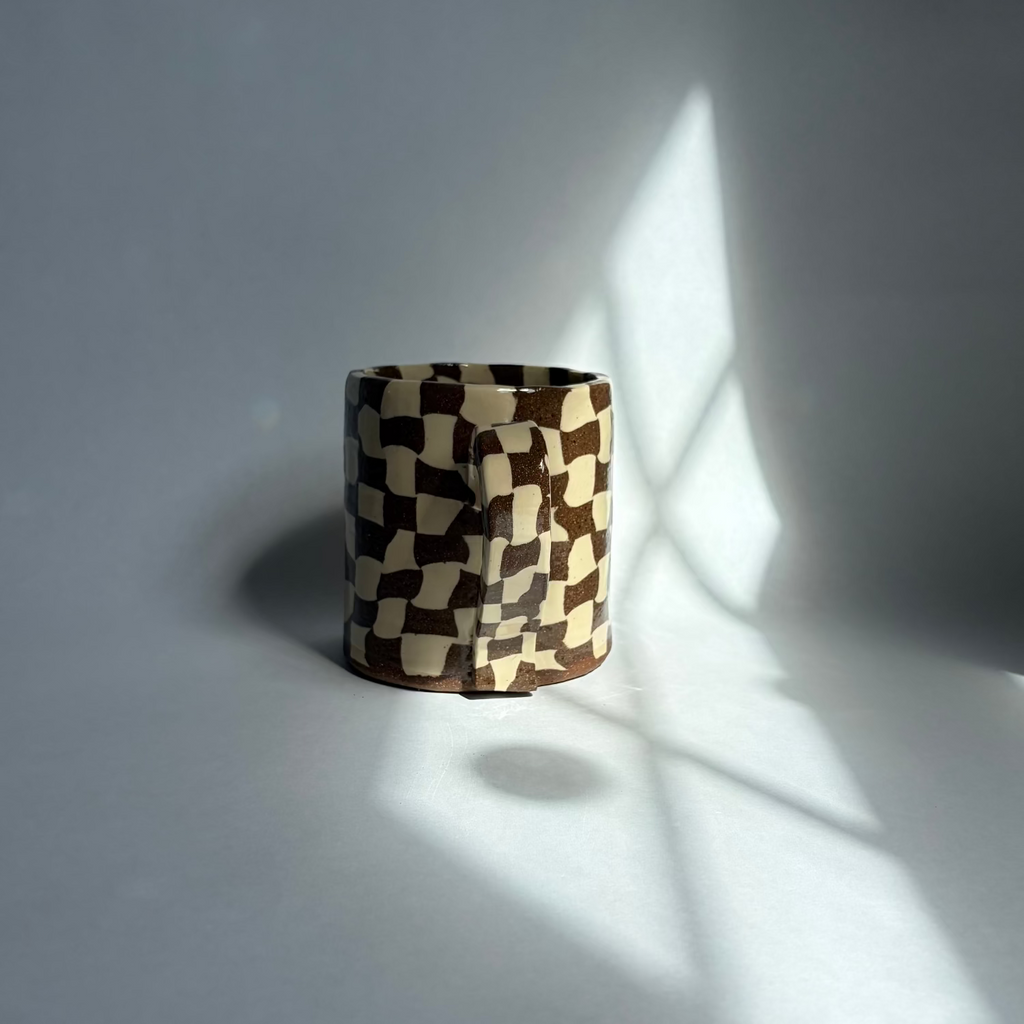 A small cylindrical product with a brown and cream checkerboard pattern, standing on a light gray surface with dramatic window-light shadows cast across the scene.