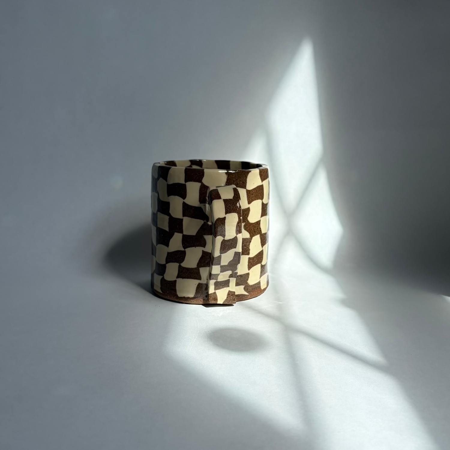 A small cylindrical product with a brown and cream checkerboard pattern, standing on a light gray surface with dramatic window-light shadows cast across the scene.
