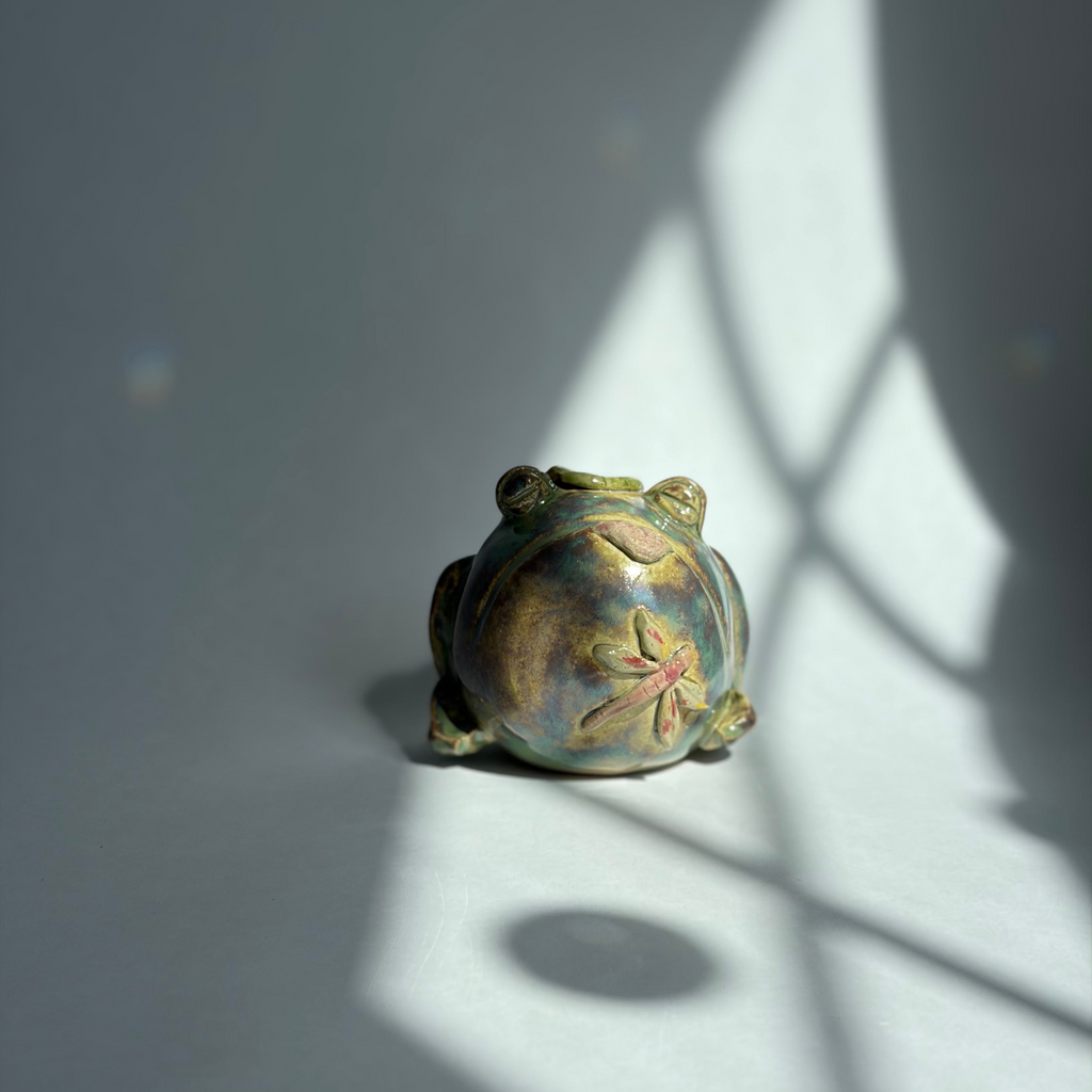 A small decorative frog figurine with a metallic gold and green finish and a pink floral detail on its back, photographed on a gray surface with dramatic window shadow patterns.