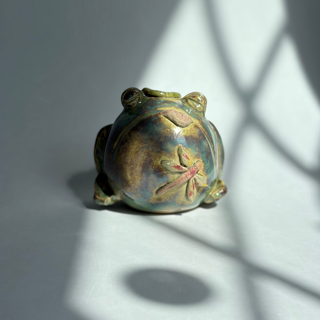 A small decorative frog figurine with a metallic gold and green finish and a pink floral detail on its back, photographed on a gray surface with dramatic window-light shadows.