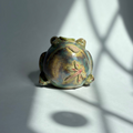 A small decorative frog figurine with a metallic gold and green finish and a pink floral detail on its back, photographed on a gray surface with dramatic window-light shadows.