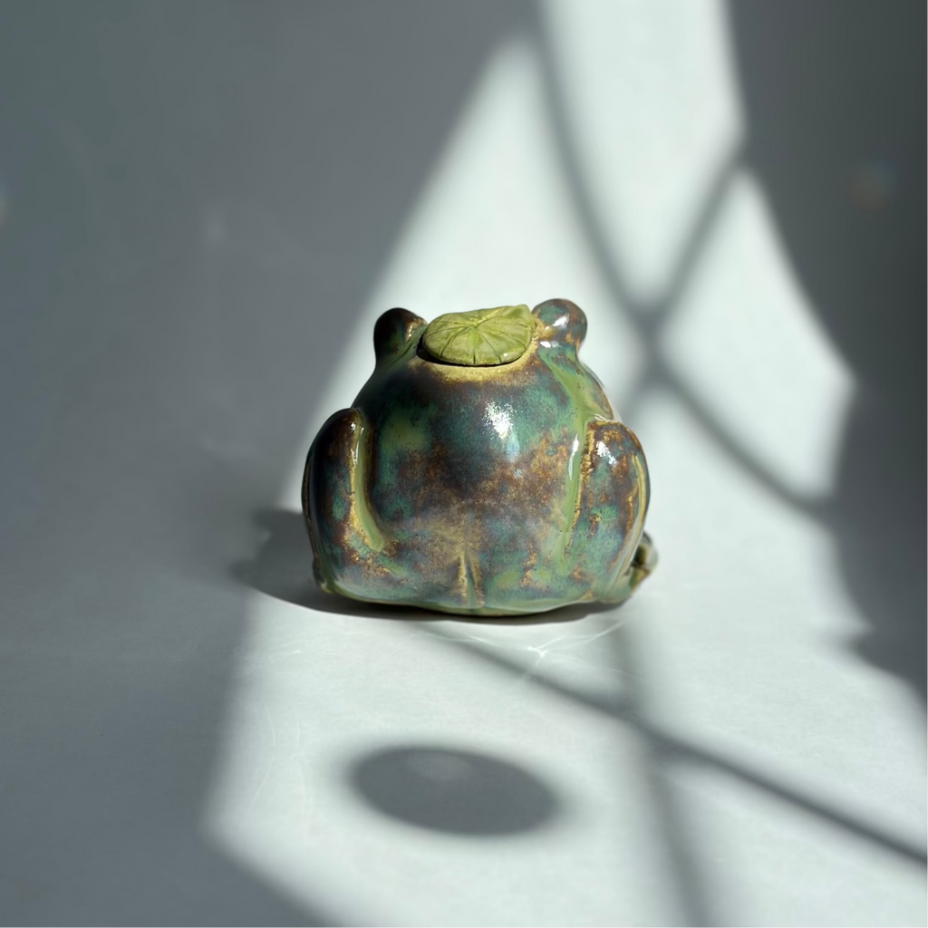 A small ceramic frog figurine with a textured, iridescent green and blue glaze, sitting on a gray surface under dramatic diagonal window shadows.