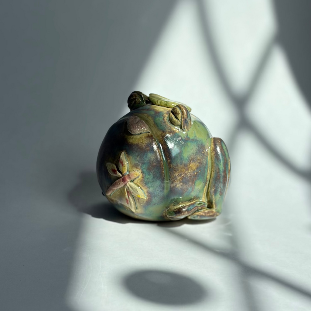A small decorative frog figurine with a glossy iridescent green and blue finish and gold-toned floral accents, photographed on a gray surface with dramatic window-light shadows.
