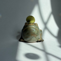 A small frog-shaped ceramic container or lidded dish with a green top, photographed on a light gray surface with dramatic window-shadow lighting.