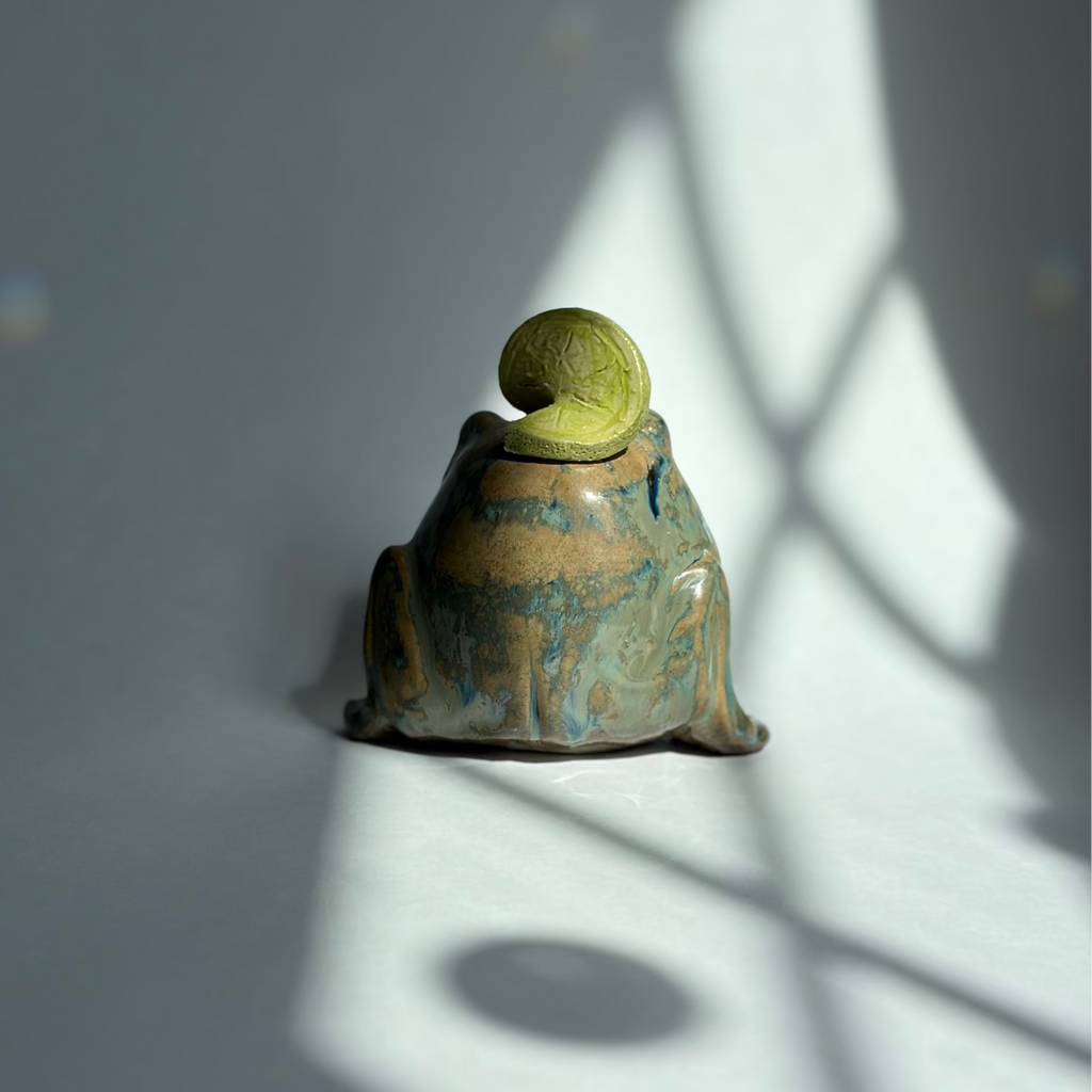 A small ceramic frog figurine with a textured green top and a blue-green glazed body, photographed on a gray surface with dramatic window shadow patterns.