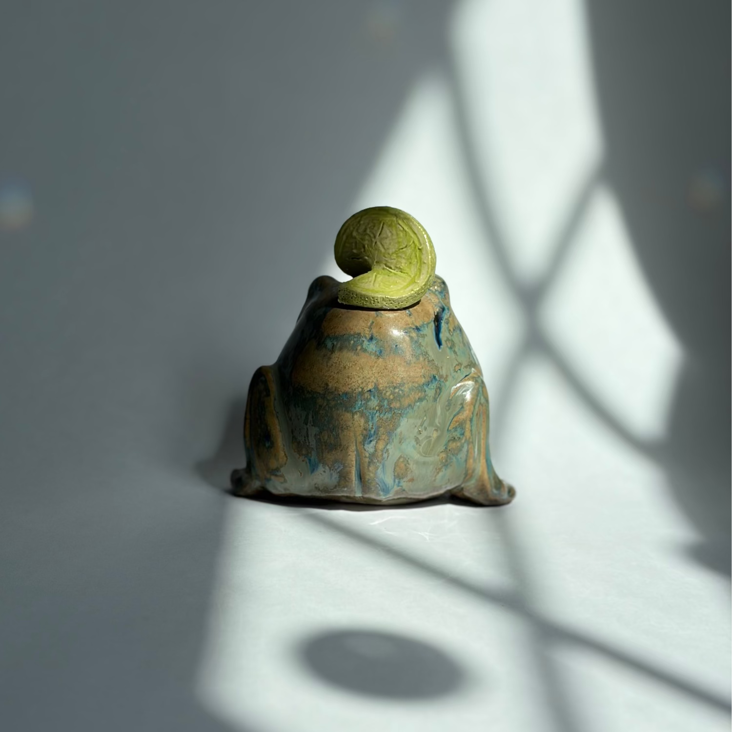 A small ceramic frog figurine with a textured green top and a blue-green glazed body, photographed on a gray surface with dramatic window shadow patterns.