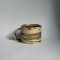 A handmade ceramic mug with a marbled pattern of beige, cream, and dark brown, set against a neutral gray studio background.