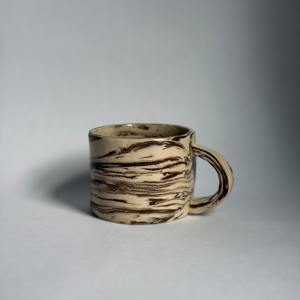 A handmade ceramic mug with a textured, wood-grain-like pattern in beige and dark brown, set against a neutral gray background.