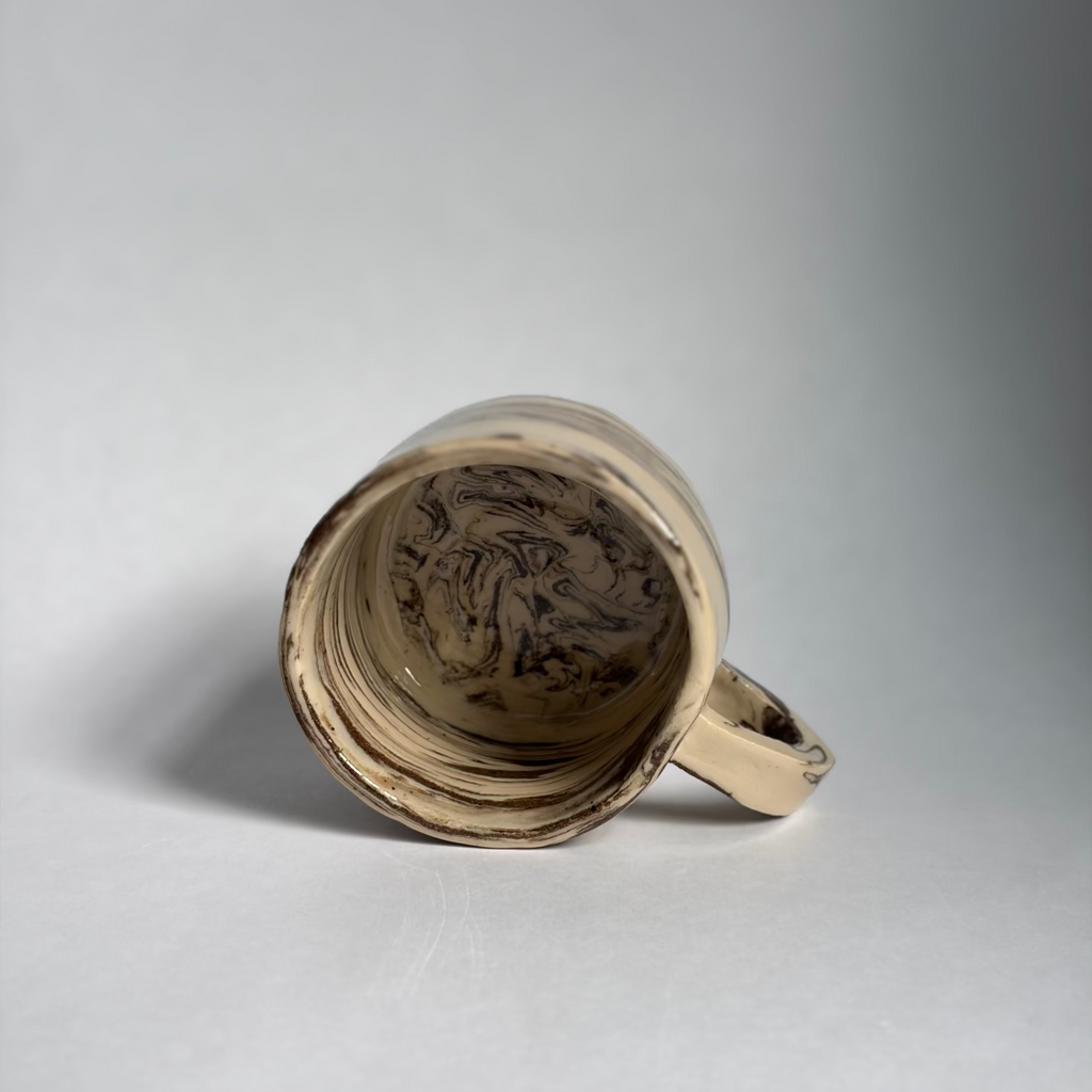 A handmade ceramic mug with a handle, shown on its side to display the interior. The mug features a natural, unglazed beige clay body with dark brown and black organic swirls and textures inside, and a smooth, matching beige exterior.