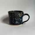 A handmade ceramic mug with a dark blue and black glaze featuring light blue and cream speckled patterns, photographed on a neutral light gray background.