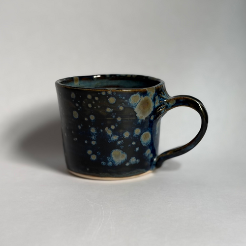 A handmade ceramic mug with a dark blue and black glaze featuring light blue and cream speckled patterns, photographed on a neutral light gray background.