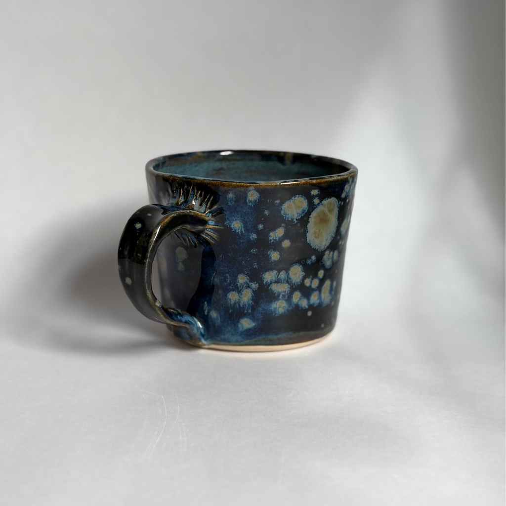 A handmade ceramic mug with a dark blue and black glaze featuring light blue and cream speckled patterns, photographed on a neutral light gray background.