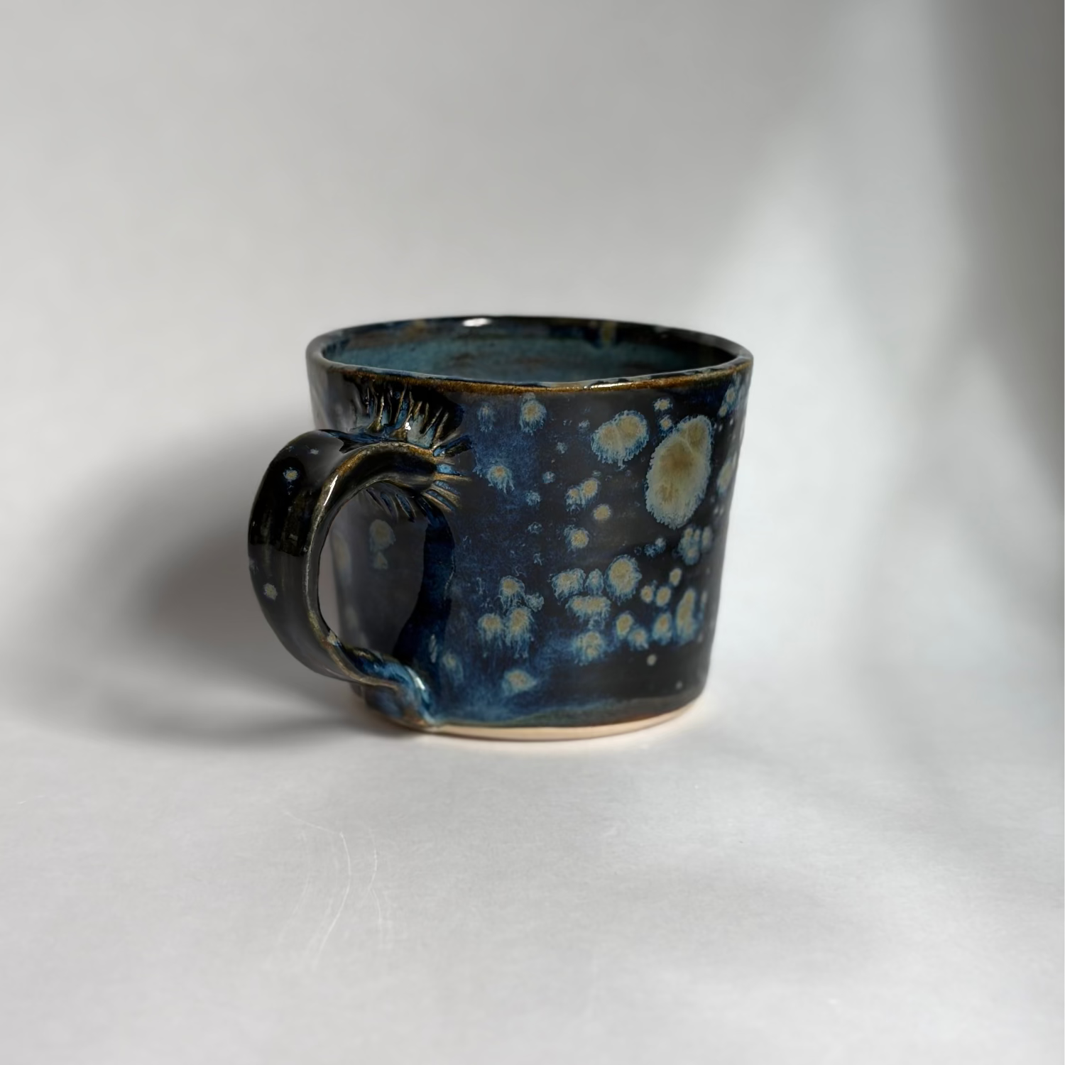 A handmade ceramic mug with a dark blue and black glaze featuring light blue and cream speckled patterns, photographed on a neutral light gray background.