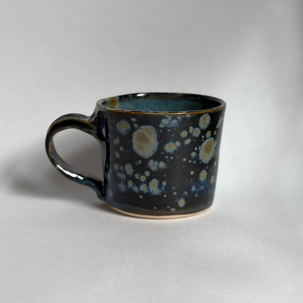 A handmade ceramic mug with a dark blue and black glaze featuring light blue and cream speckled patterns, photographed on a neutral light gray background.