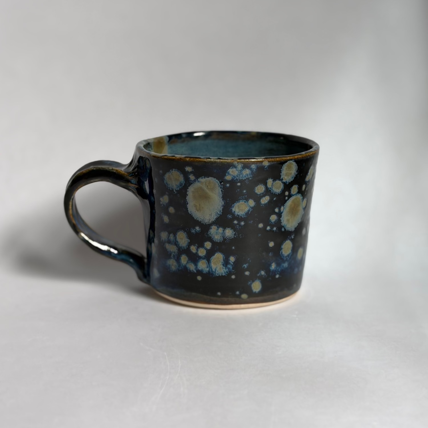 A handmade ceramic mug with a dark blue and black glaze featuring light blue and cream speckled patterns, photographed on a neutral light gray background.
