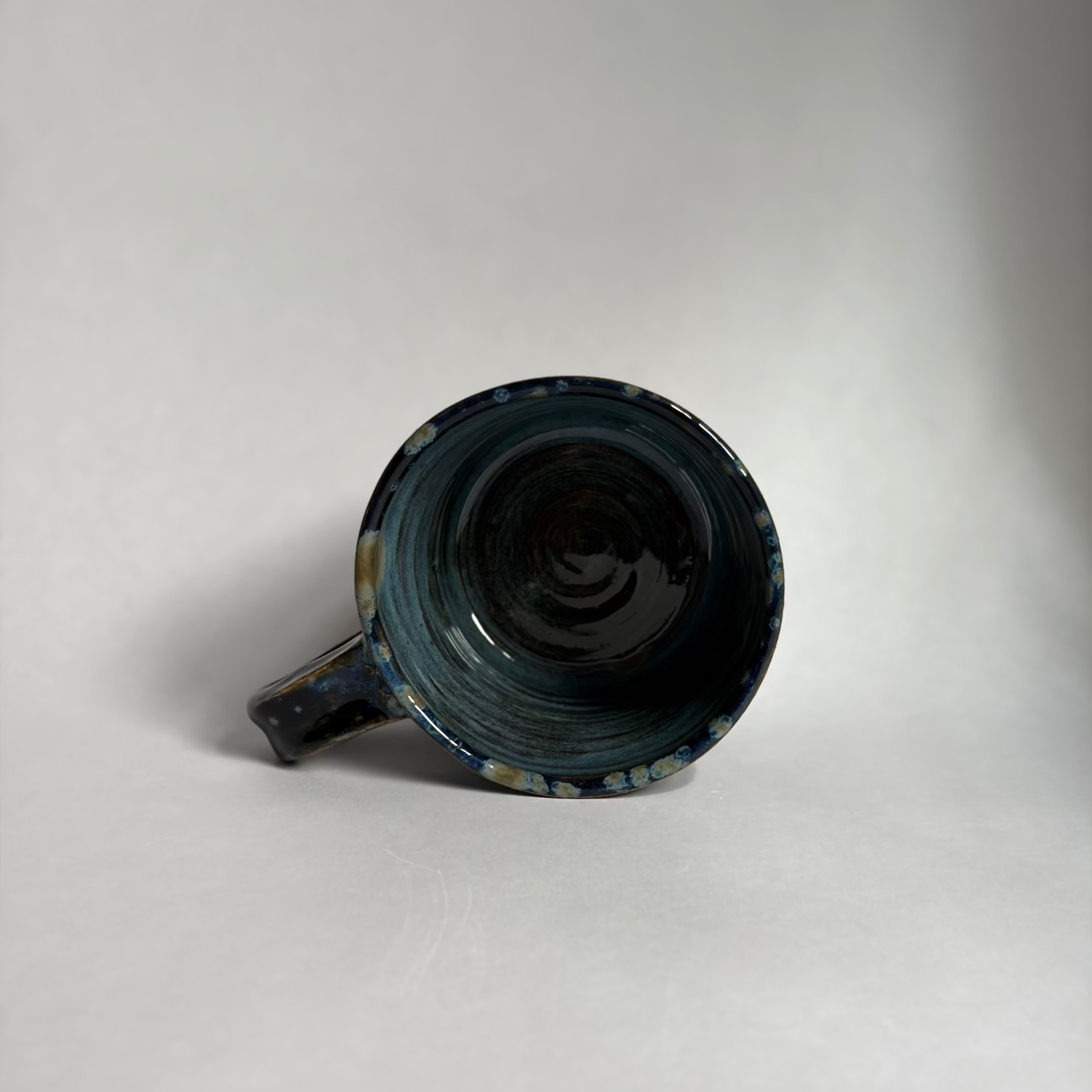 A handmade ceramic mug with a dark blue and black glaze and subtle gold speckles, shown from an overhead angle on a neutral light gray background.