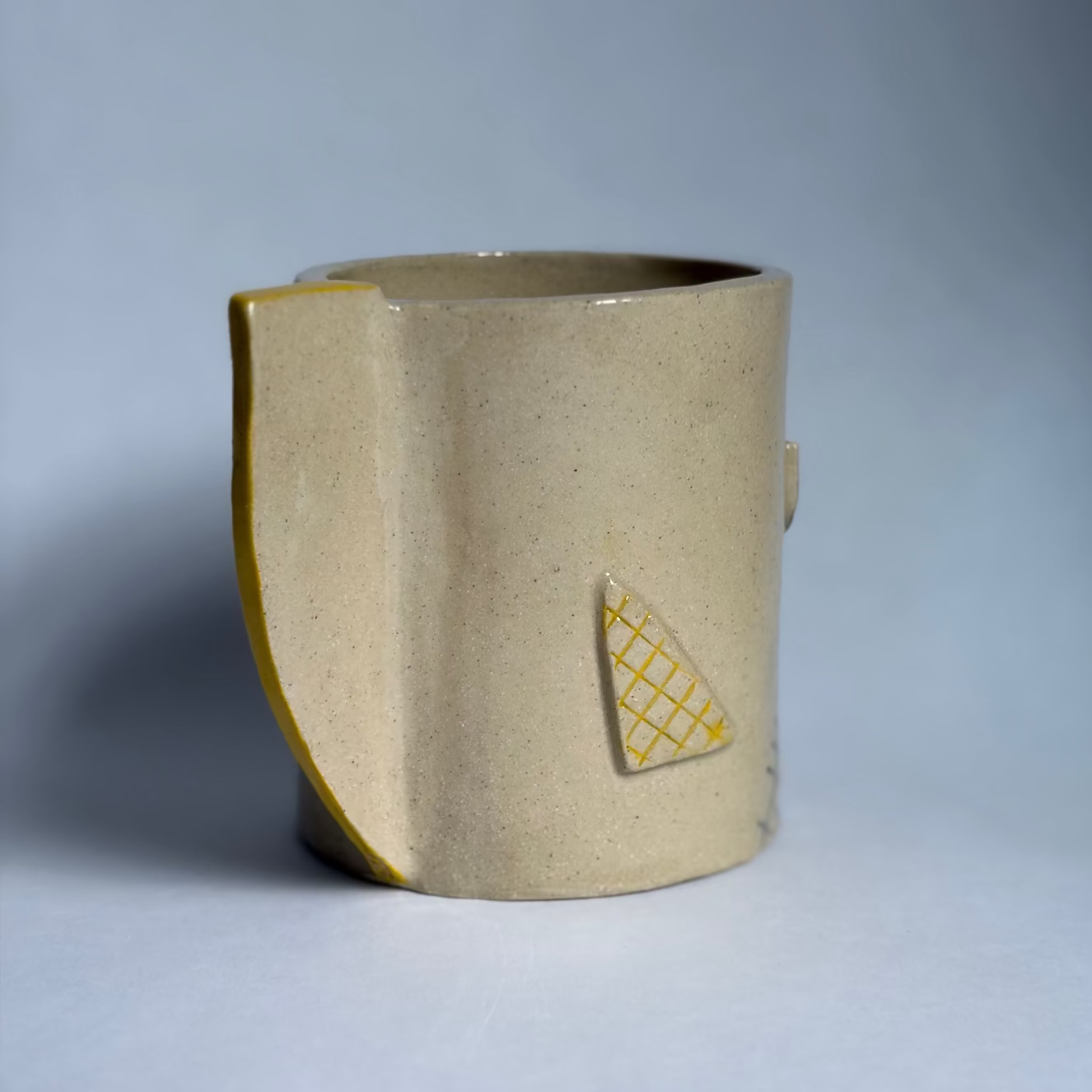 A handmade ceramic mug with a textured beige body, a yellow-gold glazed rim, and a small yellow geometric pattern on the side, set against a neutral gray background.