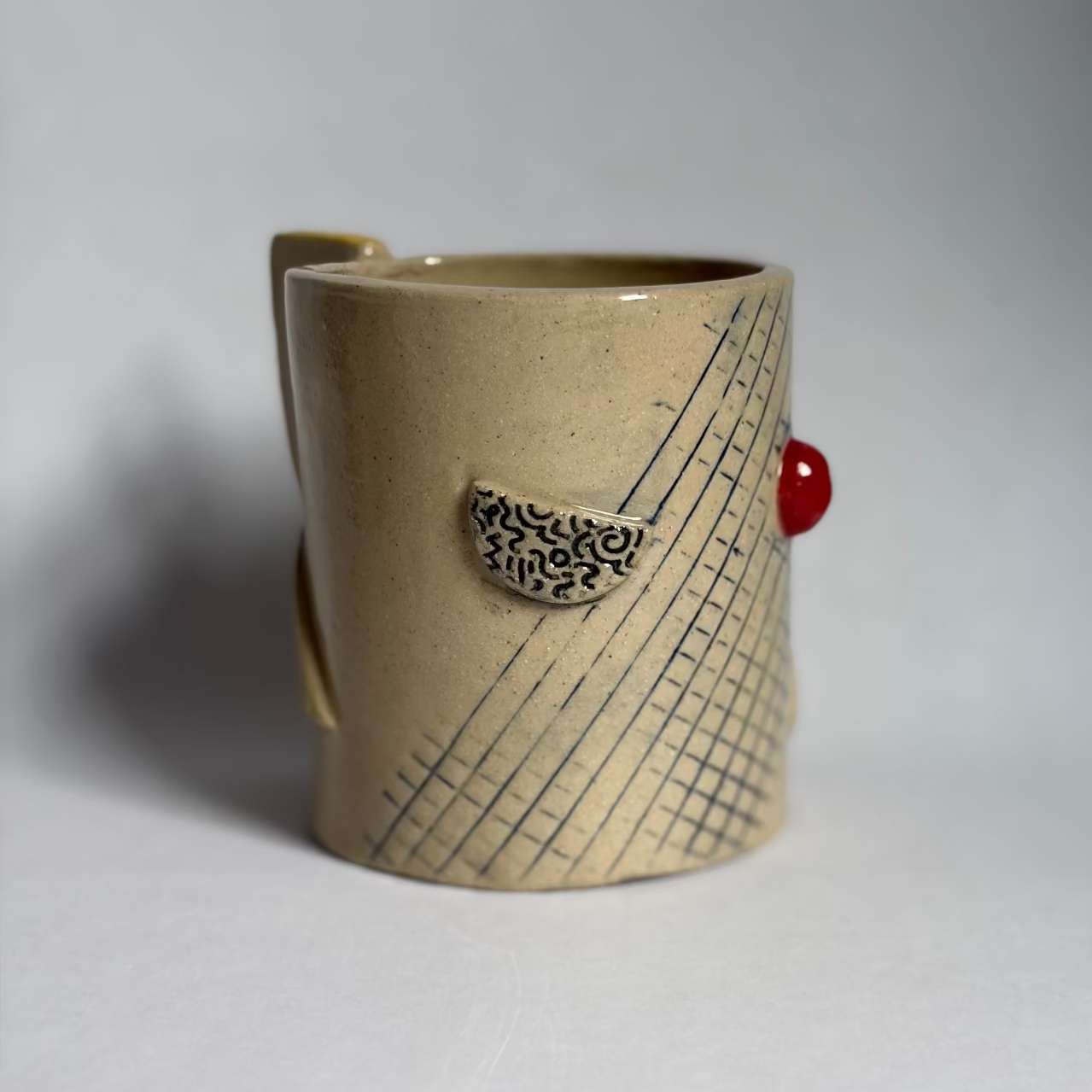 A handmade ceramic mug with a textured beige body, a yellow-gold glazed rim, and a small yellow geometric pattern on the side, set against a neutral gray background.