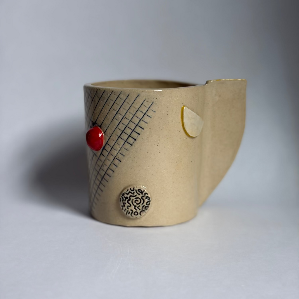 A handmade ceramic mug with a textured beige body, a yellow-gold glazed rim, and a small yellow geometric pattern on the side, set against a neutral gray background.