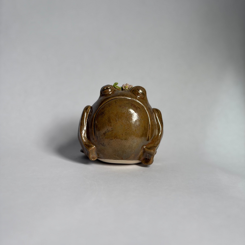 A small glazed ceramic frog figurine shown from the back, featuring a glossy brown and amber-brown finish with a subtle light-colored base, set against a neutral gray studio background.