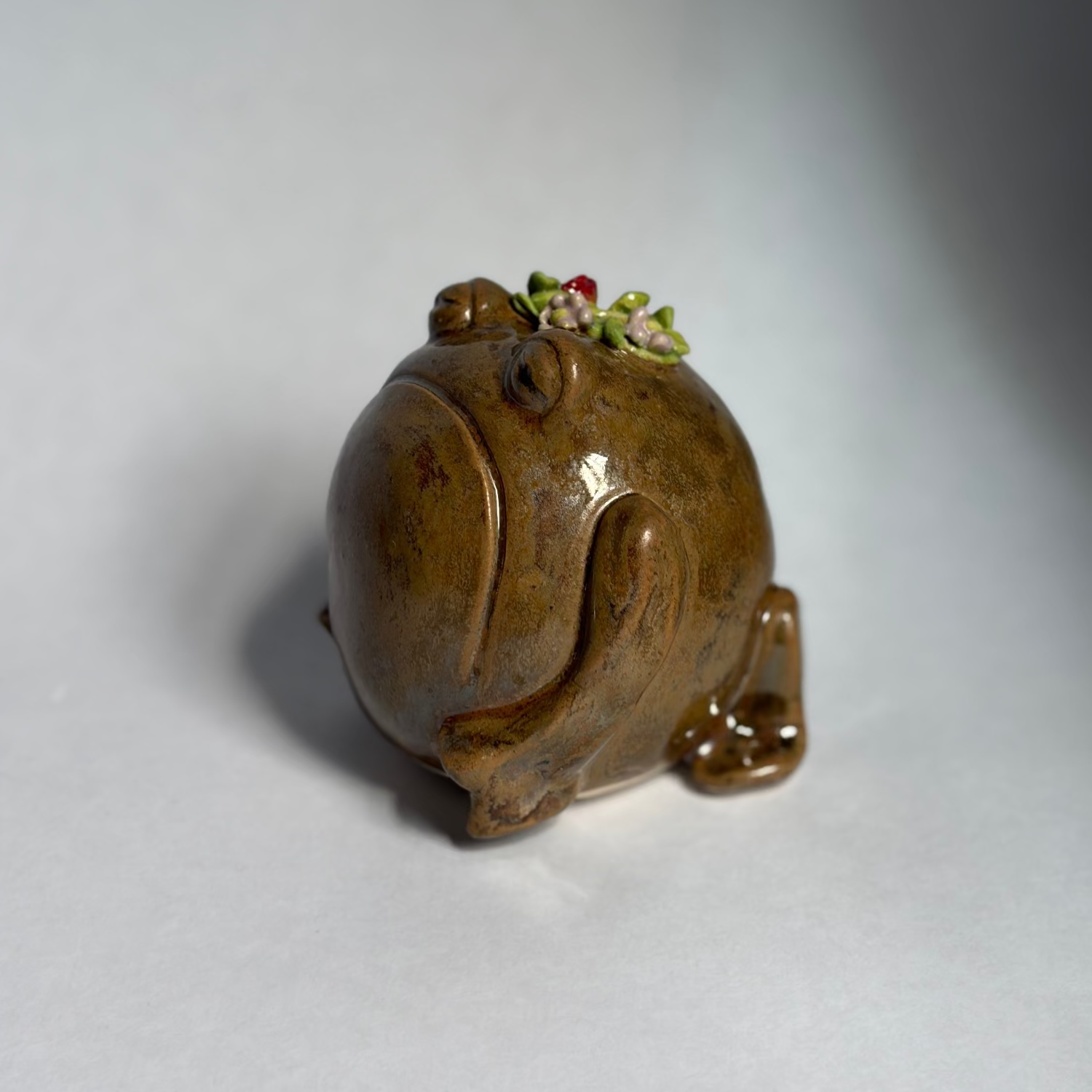 A small glazed ceramic frog figurine in earthy brown and amber tones, with tiny green and red floral accents on its head, photographed on a plain light gray background.