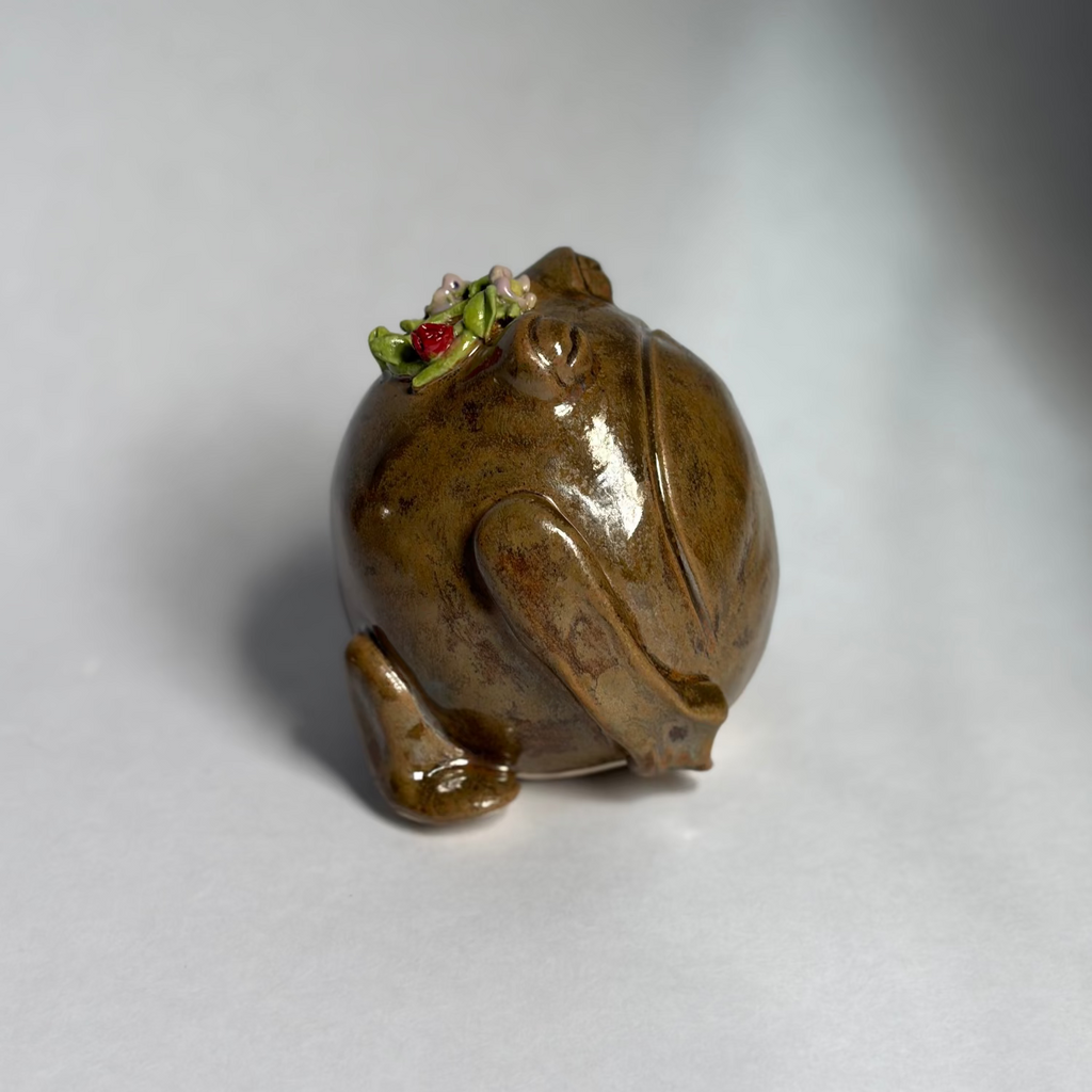 A small glossy ceramic frog figurine in earthy brown and olive tones, with tiny green and red floral accents on its head, photographed on a plain light gray background.
