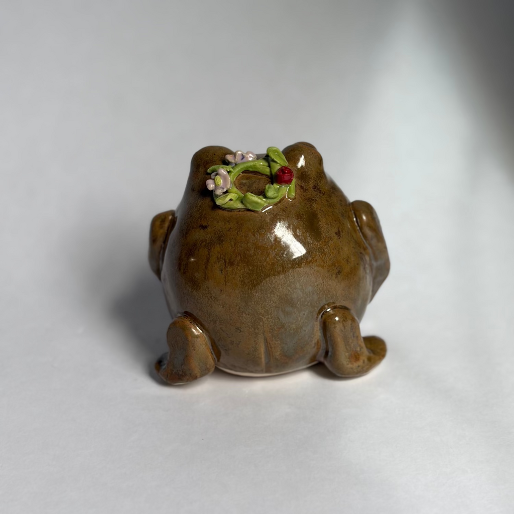 A small glossy brown frog-shaped container or planter with a green and white floral detail on its back, photographed on a plain light gray to white studio background.