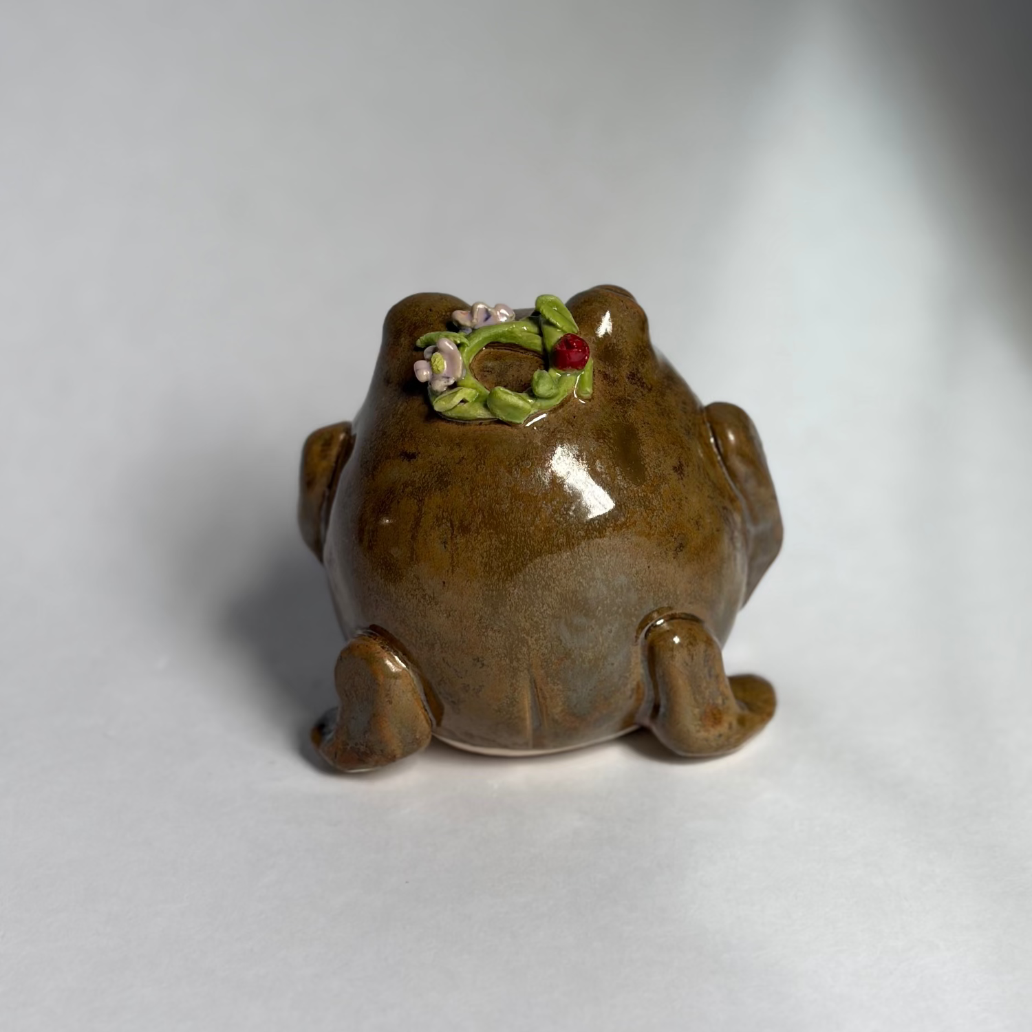 A small glossy brown frog-shaped container or planter with a green and white floral detail on its back, photographed on a plain light gray to white studio background.